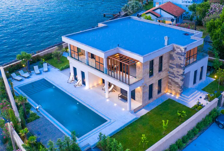 Overhead view of the villa: lit terrace, lap pool, garden and flat roof reading as one plan beside deep water.
