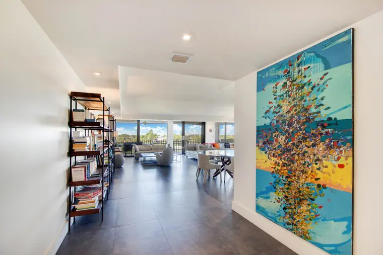 A living room with floor-to-ceiling glazing, a tall artwork and open shelving above a pale stone floor.